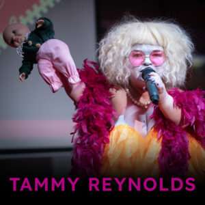 Photograph: A head and shoulders close up of a very pale skinned person with white blond curly hair. They hold a microphone to their mouth with their hand on the right. They are wearing white face paint, pink tinted glasses, a pearl necklace and yellow top with a pink feather boa around their shoulders. On top of the photograph is the text 'Tammy Reynolds' in purple lettering.