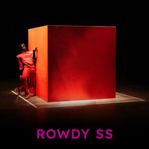 Photograph: A large orange cube in front of a black background with a figure standing, leaning on the left side of the cube. The cube is a bit taller than the person. They are a Black person dressed in orange, leaning their shoulder on the cube with their body facing us, tilting backwards. Their hand is touching the cube. On top of the photograph is the text 'Rowdy SS' in purple lettering.