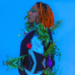 Ray Young is turned to the left, side profile. They are on an over-saturated turquoise background and they wear a fuzzy turquoise and navy jumper with a geometric design. They have plants and flowers attached to their clothes and their orange dreadlocks almost glow. The bottom of the picture reflects on itself, as if Ray is stood in water.