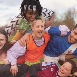 The cast of Pitch are outside in a field against a grey sky with a hedge in the background. They are climbing on each other and grinning and they all wear brightly coloured sportswear.