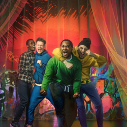 The cast of One Way Out are on a stage, leaning towards the camera. They look like they are boisterously celebrating - they have their mouths open as if they are shouting and their arms around each other. They are against a brightly coloured background with multi-coloured graffiti and sheer, pale yellow mesh curtains to either side.
