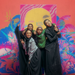 The cast of Dugsi Dayz are standing huddled together in the centre of the photo, posing and smiling or pulling fun faces. They stand in front of a bright, multicoloured background with artwork of painted flowers, plants and clouds, and chalk drawings of animals and other doodles.