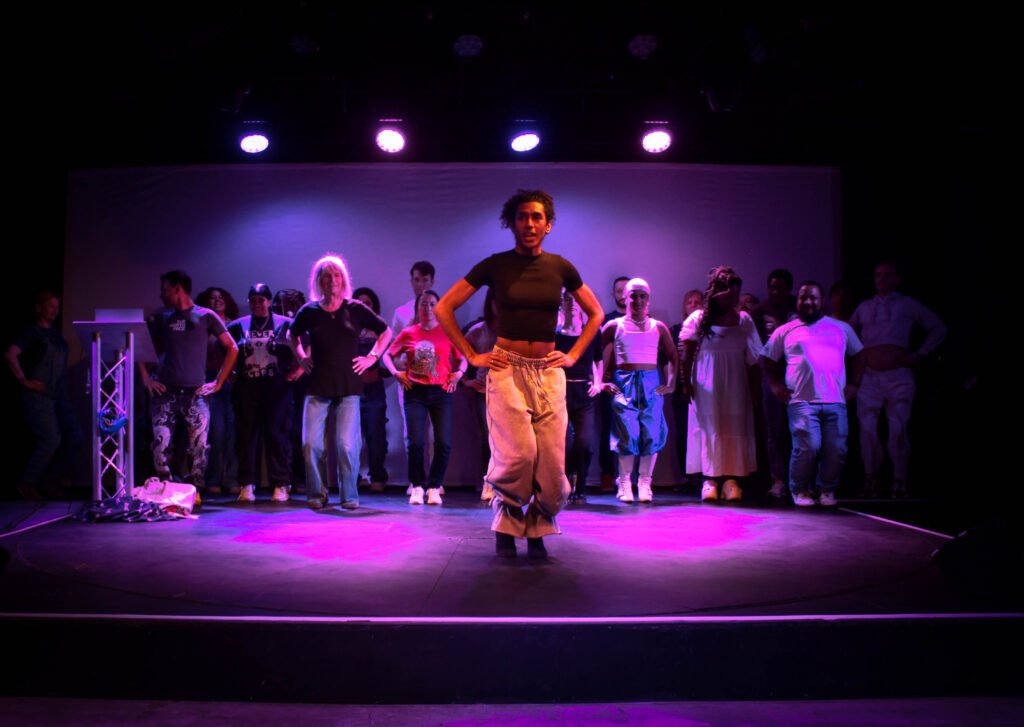 A group of workshop participants stand behind an instructor (Bambi) on a purple lit stage, with their hands on their hips.