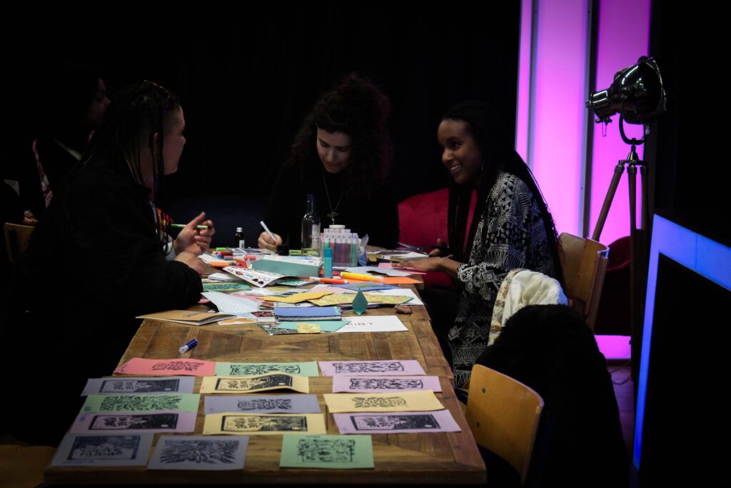 Three people sit around a table while drawing and cutting paper, smiling and talking.