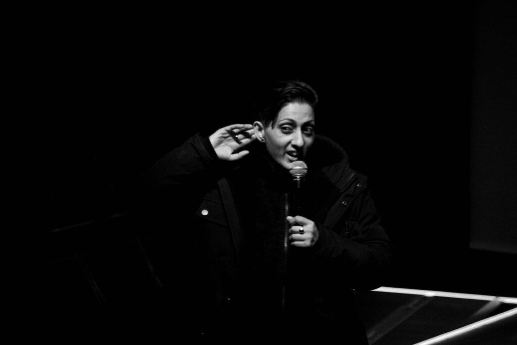 A performer stands on stage in black and white, speaking into a microphone and motioning to their ear.
