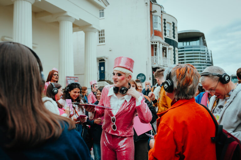 Alfie Ordinary, a drag prince dressed in pink sequins, is taking a group of people on a walking tour. Alfie is holding onto a headset, and the people around him are all wearing wireless headphones and getting ready to go on the tour.
