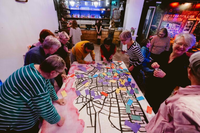 Nine women are gathered around a large canvas map of Brighton and Hove. The women are pinning memories of their lives onto the map, with colorful tags.